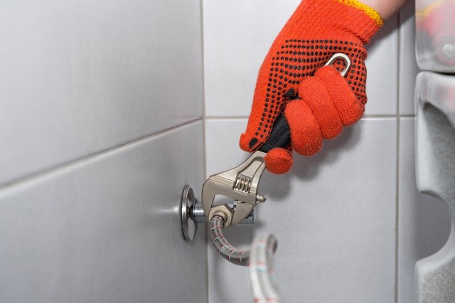 a homeowner in orange gloves tightening a braided metal hose on a toilet