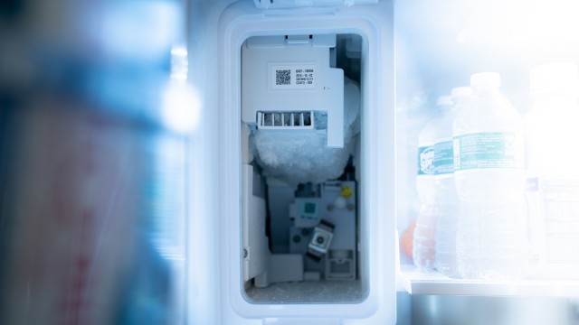 The ice maker in a freezer is currently without its bin, which shows its mechanical parts